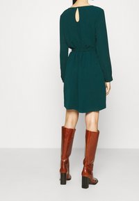 Turkusowa sukienka z długimi rękawami i detalem w kształcie dziurki z tyłu, taliowana w pasie. Połączona z brązowymi skórzanymi botkami za kolano na kwadratowych obcasach.