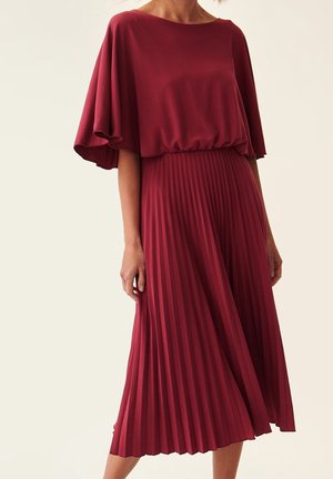 Donna che indossa un vestito color bordeaux con una parte superiore larga e una gonna plissettata, in piedi contro uno sfondo semplice.
