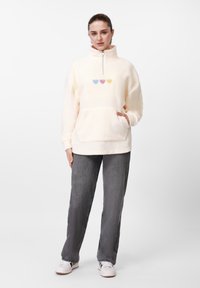 Sudadera de forro polar amarillo claro con media cremallera y gran bolsillo frontal, con tres pequeños corazones bordados en azul, rosa y amarillo.