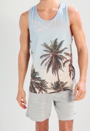 Homme portant un débardeur sans manches avec un motif de palmier et un short gris clair, se tenant devant un fond uni.