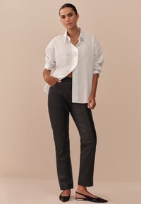 Chemise blanche à boutons avec manches retroussées, associée à un pantalon en cuir noir élégant. La tenue est complétée par des ballerines noires et un collier en or.