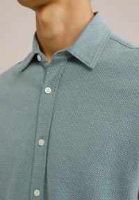 Šviesiai mėlynas marškinėlių mygtukų užsegimas su tekstūrine deimanto raštu, minkšta medžiaga ir balti plastikiniai mygtukai ant mygtukų ir apykaklės.