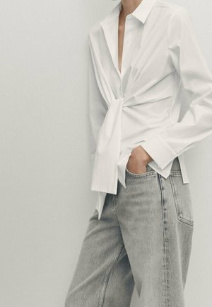 White long-sleeve shirt with a front knot paired with light gray loose-fit jeans, one hand tucked into a pocket, against a plain light background.