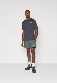 Grey short-sleeve t-shirt with white logo, paired with green shorts. Model wears black sneakers and white socks, standing against a plain background.