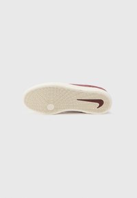 Røde semskede sneakers med en teksturert hvit gummisåle, som har en kontrasterende brun logoakse og skrågrep på bunnen.