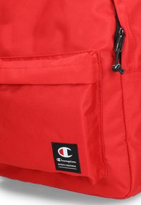 Czerwony plecak z nylonu z przednią kieszenią, czarnym zamkiem błyskawicznym i naszywką z logo Champion. Charakteryzuje się teksturowaną powierzchnią i wytrzymałym materiałem.