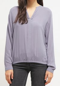 Blouse à manches longues en tissu gris clair, dotée d'un col en V, de plis à l'avant et d'un ourlet élastique, avec une texture lisse et légèrement brillante.