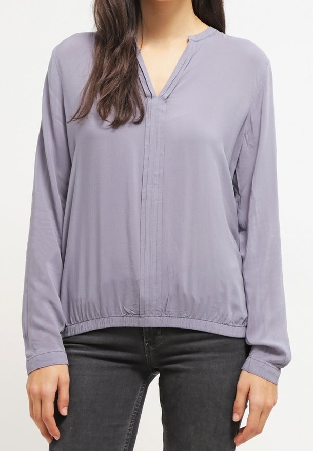 Blouse à manches longues en tissu gris clair, dotée d'un col en V, de plis à l'avant et d'un ourlet élastique, avec une texture lisse et légèrement brillante.