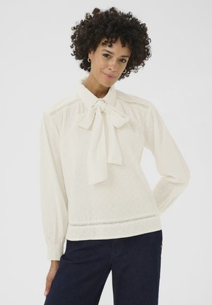 White blouse with a bow tie collar, textured fabric, long sleeves, and lace detailing along the hem, paired with dark blue jeans.