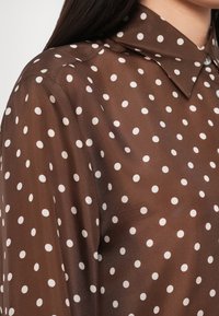 Bluza maro cu buline albe, cu o textură netedă, având guler ascuțit și detalii cu nasturi. Prim-plan al zonei umerilor.