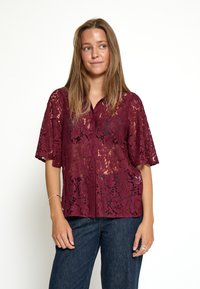 Blusa di pizzo bordeaux con chiusura a bottoni sul davanti, maniche corte e motivi floreali. Abbinata a pantaloni di denim scuri.