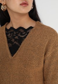 Maglione marrone a scollo a V con una finitura testurizzata, indossato sopra pizzo nero. Orecchini a cerchio dorati aggiungono contrasto.