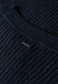 Navy knitted sweater with textured fabric, featuring a ribbed collar and a prominent black metal logo tag labeled "SUPERDRY."