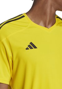 Geel sportshirt met V-hals, voorzien van zwarte drie-streep accenten op de schouders en een zwart Adidas-logo op de borst.