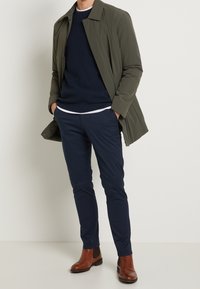 Mann in olivgrünem Mantel, mit marineblauem Pullover über einem weißen Hemd, marineblauen Hosen und braunen Leder-Chelsea-Stiefeln, der mit den Händen in den Taschen dasteht.