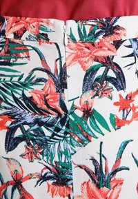 Tissu blanc avec un motif floral vibrant en rouge, vert et bleu, présentant de grandes fleurs tropicales et des feuilles, avec une fermeture éclair.