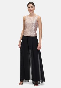 Un débardeur en sequins rose gold associé à un pantalon large noir en tissu transparent, avec des plis verticaux et une silhouette fluide.