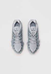 Αθλητικά παπούτσια Nike Shox με γκρι πλέγμα πάνω μέρος, ριγωτό σχέδιο και μοναδική μαξιλαρωτή σόλα. Οι λεπτομέρειες περιλαμβάνουν σκούρες γκρι ρίγες και λογότυπα.