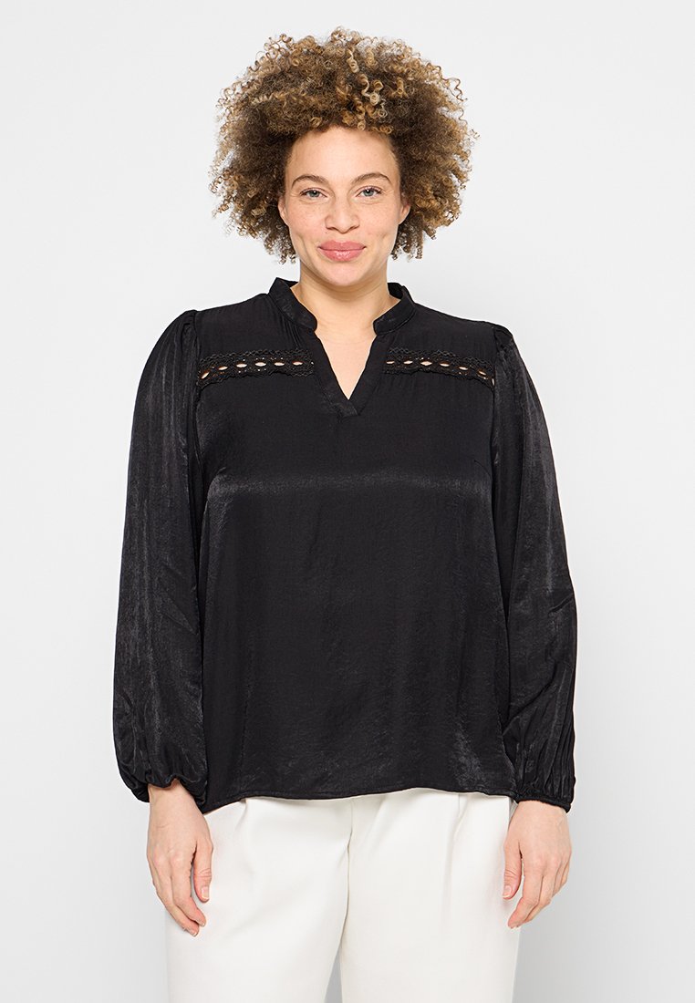 Blouse noire à manches longues, décolleté en V et accents découpés décoratifs sur le plastron. Texture lisse avec une coupe décontractée.