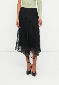 Saia preta de renda com uma barra assimétrica e fluida, apresentando um padrão floral e um design em camadas. Combinada com sapatos de salto pretos.