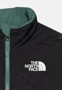 Svart jacka med en grön inre krage, utrustad med en fullständig dragkedja och en vit logotyp på vänster bröst. Materialet verkar slätt och syntetiskt.