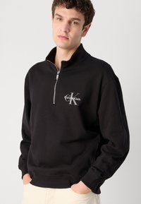Musta huppari, jossa on vetoketjullinen kaulus ja harmaa "Calvin Klein" -logo. Pehmeä kangas, resorit hihansuissa ja helmassa, rento istuvuus.
