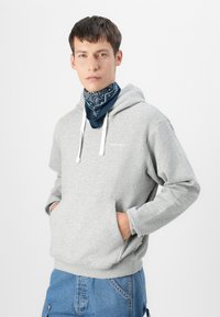 Sudadera gris con capucha, cuerdas blancas y bolsillo tipo canguro. El modelo lleva una bandana azul marino alrededor del cuello con patrones blancos.