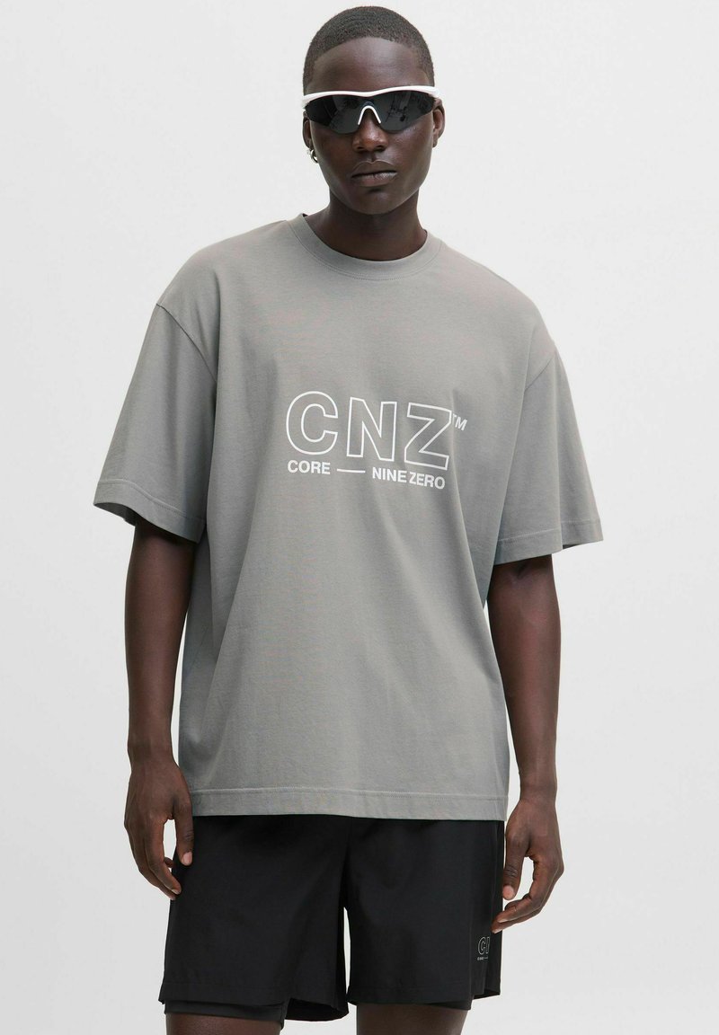 Grå oversized t-shirt med hvid "CNZ," parret med sorte shorts. T-shirten har korte ærmer og en rund hals. Sorte solbriller fuldender looket.