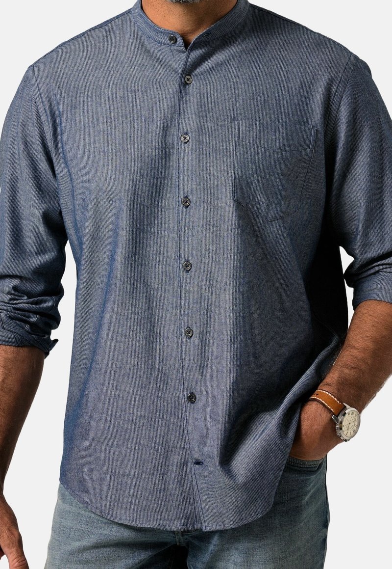 Homme portant une chemise bleue à manches longues avec col mandarin, manches retroussées, jean en denim et une montre-bracelet avec un bracelet en cuir marron.