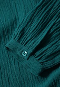 Teal skjorta med samlade ärmar som har texturerad tyg, synliga veck och en kontrasterande blå knapp vid manschetten.