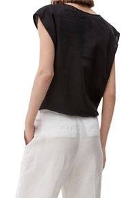 Donna che indossa una canottiera nera senza maniche e pantaloni bianchi larghi, in piedi con una mano in tasca, vista da dietro.