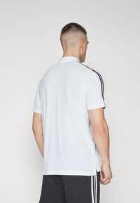 Hvit polo skjorte med korte ermer, med svarte striper langs skuldrene. Materialet virker mykt, med en klassisk krage og rett kant.