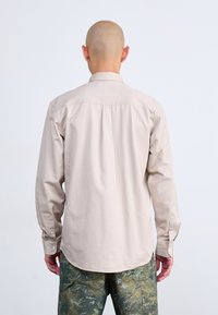 Camisa abotonada de un color beige claro con un detalle de yugo trasero, mangas largas y un dobladillo curvado, combinada con pantalones cargo verdes con estampado.
