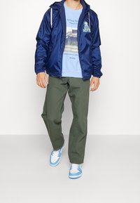 Marinkapp med huva, tryckt ljusblå t-shirt, olivgröna byxor och blå sneakers med vita detaljer. Enkelt mönster med dragsko.