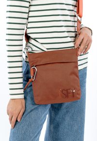 Brūna pinuma pārvietojamā soma ar teksturētu virsmu, rāvējslēdzēja aizdari un metāla aizdares detaļu, attēlota pret svītrainas krekla fonu.