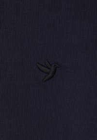 Marine stof med en struktureret overflade, der har et lille, sort broderet kolibri-logo i nederste venstre hjørne.