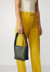 Groene leren tas met een rechthoekige vorm, korte zwarte handgreep en subtiele stiksels, vastgehouden door een persoon in gele broek.