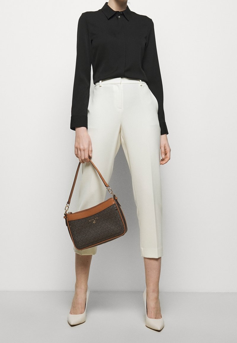 Femme portant une chemise noire à manches longues, pantalon blanc court, talons blancs, tenant un sac à main marron à motif sombre à côté.
