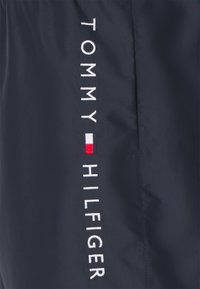 Veste marine avec le texte brodé "TOMMY HILFIGER" en blanc, accompagné d'un petit emblème en rouge, blanc et bleu. Tissu lisse et léger.