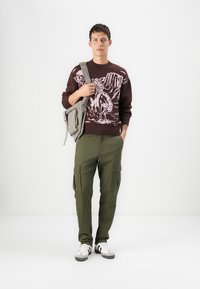 Suéter marrón de punto con diseño gráfico blanco, pantalones cargo verdes, bolso de hombro gris y zapatillas blancas con detalles negros.