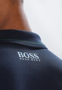 Närbild av en persons nacke som bär en marinblå polo med "BOSS HUGO BOSS" tryckt under kragen och med gröna sömdetaljer.