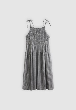 Robe sans manches à carreaux vichy noir et blanc avec corsage smocké et bretelles réglables nouées, jupe de longueur mi-mollet, sur fond clair uni.