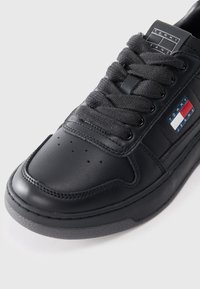 Black leather sneaker with textured gray laces, perforated toe, gray sole, and a side label featuring red, white, and blue accents.