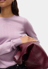 Maglione in lana viola chiaro con polsini a coste, indossato con una borsa in pelle bordeaux scuro. Texture liscia, scollo rotondo semplice, dettagli minimi.