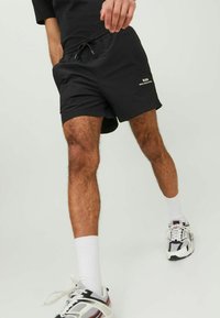 Schwarze Shorts mit einem elastischen Bund und Kordelzug. Mit Seitentaschen und einem kleinen weißen Logo am Saum. Getragen mit Sportschuhen und Socken.