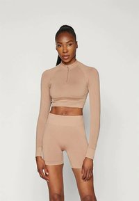 Frau mit langen Haaren in einem hohen Pferdeschwanz, die ein beiges, strukturiertes Langarm-Crop-Top und dazu passende hochgeschnittene Radlerhosen trägt und aufrecht steht.