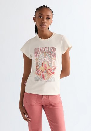 Νεαρή γυναίκα με πλεγμένα μαλλιά που φοράει λευκό μπλουζάκι Wrangler με σχέδιο και ροζ παντελόνια, στέκεται μπροστά σε απλό φωτεινό φόντο.