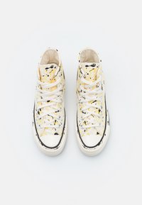 Baskets montantes blanches avec un design de taches de peinture jaunes et noires, lacets blancs et semelles en caoutchouc sur un fond blanc uni.