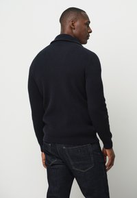 Ciemnoniebieski sweter z prążkowanej dzianiny z złożonym kołnierzem, dopasowany fason i długie rękawy, zestawiony z ciemnymi dżinsami.