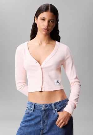 Junge Frau mit langen dunklen Haaren, die eine hellrosa kurz geschnittene Strickjacke mit Knöpfen und blaue, hoch taillierte Jeans vor einem grauen Hintergrund trägt.